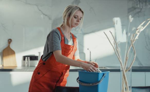 Home A woman in a gray shirt and red overalls cleans a kitchen counter using a blue bucket and white rag. Modern setting.